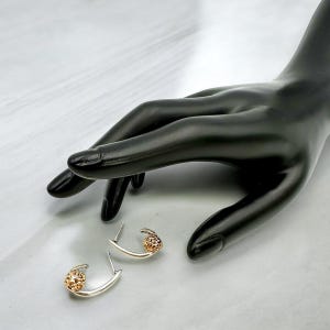May include: A pair of gold-toned earrings with a unique design. Each earring features a curved, silver-colored bar with a textured, gold-colored sphere at the end. The earrings are displayed on a black hand against a white background.