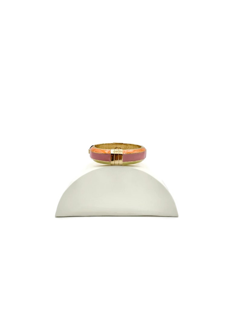 May include: A gold ring with a pink and orange enamel band. The ring is sitting on a white display stand.