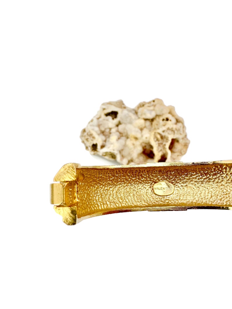 May include: Gold-toned metal bracelet with a textured surface and a circular logo that reads "VALENTINO" on the inside.