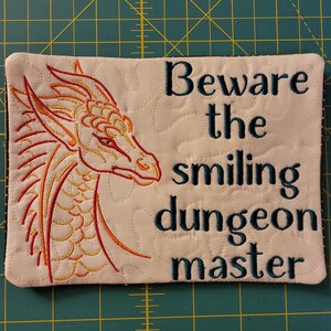 May include: A beige fabric coaster with an embroidered orange dragon head on the left and the text "Beware the smiling dungeon master" in dark blue on the right.