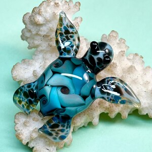 Peut inclure: Une figurine de tortue de mer en verre présentant un motif tourbillonnant de couleurs bleu profond, turquoise et noir. La tortue a une finition lisse et brillante avec des taches sombres distinctes sur ses nageoires et sa tête.