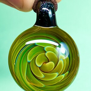 Peut inclure: Un pendentif en verre vert avec un motif en spirale. Le pendentif est attaché à un cordon brun.