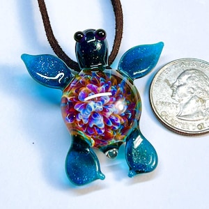 Peut inclure: Un pendentif en verre de tortue de mer bleu turquoise avec un motif floral iridescent et coloré au centre. Le pendentif est sur un cordon en cuir brun.