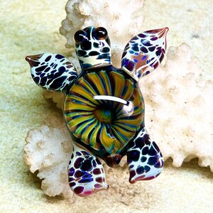 Peut inclure: Une petite figurine de tortue de mer en verre avec une carapace irisée et tourbillonnante. La tortue a une carapace tacheté de blanc et de noir avec des accents de violet et de bleu. Les yeux de la tortue sont bruns et noirs.