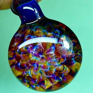 Peut inclure: Un pendentif en verre artisanal avec un motif tourbillonnant vibrant. Le pendentif présente un dessus bleu foncé qui se transforme en un globe rond et transparent rempli de motifs colorés de jaune, orange, violet et bleu. Le verre est suspendu à un cordon noir.