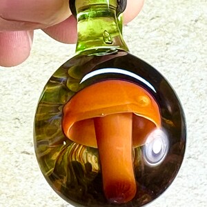 Peut inclure: Un pendentif en verre avec un dessus vert clair et un fond rond et clair. À l'intérieur du pendentif se trouve un champignon en verre avec un chapeau orange et un pied blanc.