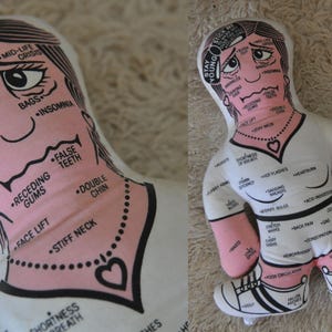 May include: A novelty pillow in the shape of a person, with a white body and pink limbs. The pillow features a diagram of the body with labels for various ailments, including wrinkles, insomnia, and hot flashes. The text "Stay Young" is printed on the head.