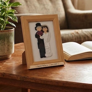 May include: A framed beaded wedding artwork showing a bride and groom. The artwork is in a light wood frame, displayed on a wooden table. The bride is in a white dress, and the groom wears a black suit. The text "Beaded Perfectly Carol Webb" is at the bottom.