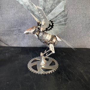 May include: A sculpture of a bird constructed from recycled metal parts, including gears, bolts, and sheet metal. The bird features outstretched wings made of thin metal plates, a mechanical body, and stands on a circular gear base.