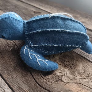 May include: A handmade blue felt turtle with white stitched details. The turtle has a rounded shape with a head, flippers, and a shell. The shell is decorated with white stitching. The turtle is lying on a wooden surface.