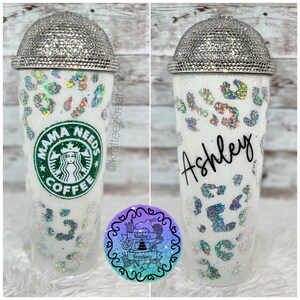 May include: A white glitter tumbler with a rhinestone-covered lid. The tumbler features a leopard print design with iridescent accents. One side displays the Starbucks logo with "Mama Needs Coffee," and the other side has the name "Ashley."