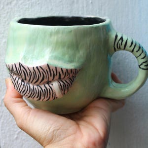 Op de afbeelding: Een lichtgroene keramische mok met een glanzende afwerking, voorzien van een gebeeldhouwd paar lippen met een zwart zebrapatroon op een lichtroze achtergrond. De mok heeft een bijpassend lichtgroen handvat versierd met zwarte zebramarkeringen.