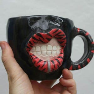 May include: Black ceramic mug with a unique design featuring a large, open mouth with red and black striped lips and white teeth. The handle also has red and black stripes. A silver barbell piercing is visible.