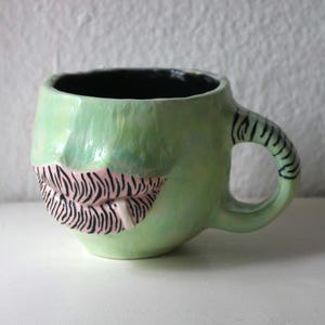 Könnte beinhalten: Ein hellgrüner Keramikbecher mit glänzender Oberfläche, der ein skulptiertes Relief von Lippen mit schwarzen, zebraartigen Streifen auf rosa Grund zeigt. Der Becher hat ein schwarzes Inneres und einen Henkel mit passenden schwarzen Streifen.