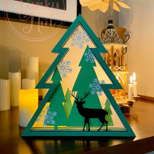 Puede incluir: Decoración en forma de árbol de Navidad verde con silueta de ciervo y recortes de copos de nieve. El árbol tiene un diseño en capas con un interior cálido y brillante. Velas y un molino de viento decorativo en el fondo.