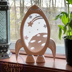 May include: White papercraft rocket ship with a moon and stars design. The rocket is sitting on a windowsill.
