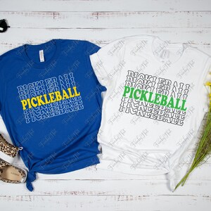 以下が含まれることがあります： 「PICKLEBALL」の文字が重ねてデザインされた、青と白の2枚のTシャツ。青いシャツは黄色のアクセント、白いシャツは緑色のアクセント。サングラスとレオパード柄の靴がアクセサリーとして添えられています。