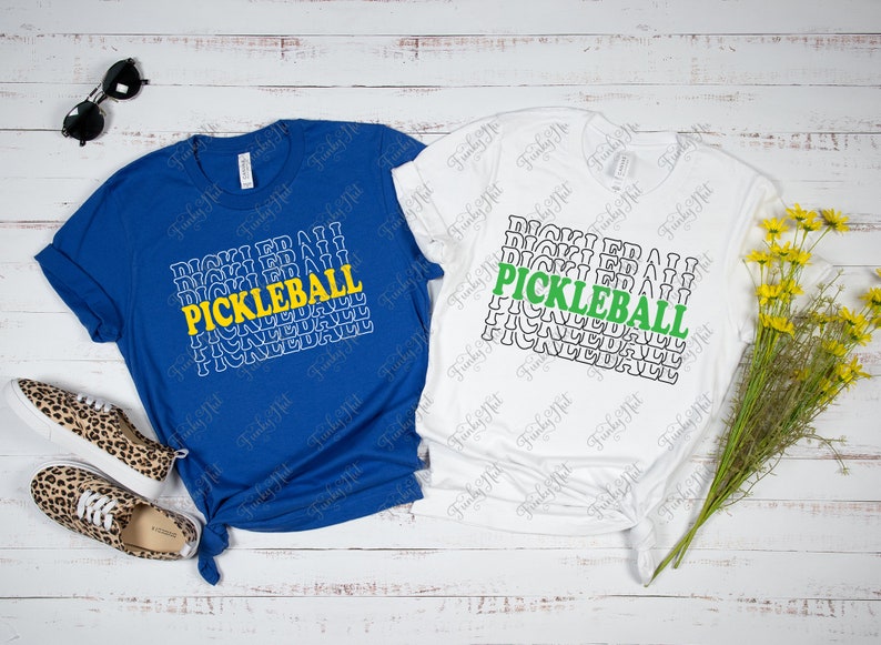 以下が含まれることがあります： 「PICKLEBALL」の文字が重ねてデザインされた、青と白の2枚のTシャツ。青いシャツは黄色のアクセント、白いシャツは緑色のアクセント。サングラスとレオパード柄の靴がアクセサリーとして添えられています。