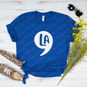 May include: A royal blue t-shirt with a white graphic of a speech bubble with the text "LA" inside. The graphic is a stylized number 9 with the letters "LA" inside the curve of the number.