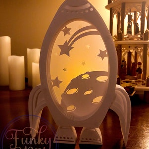 May include: A white paper rocket ship with a moon and stars cut out design. The rocket ship is lit from inside, creating a glow effect. The rocket ship is sitting on a wooden table.