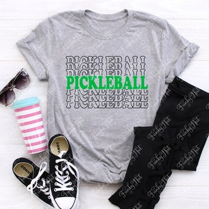 以下が含まれることがあります： 緑と黒で「PICKLEBALL」の文字が入ったヘザーグレーのTシャツ。画像には、黒のレギンス、黒のコンバーススニーカー、サングラス、ピンクと白のストライプのタンブラーも含まれています。