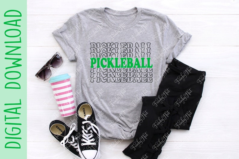 以下が含まれることがあります： 緑と黒で「PICKLEBALL」の文字が入ったヘザーグレーのTシャツ。画像には、黒のレギンス、黒のコンバーススニーカー、サングラス、ピンクと白のストライプのタンブラーも含まれています。