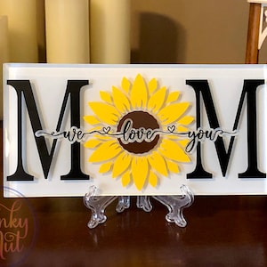 May include: White acrylic sign with black letters spelling "MOM" with a yellow sunflower in the centre. The sunflower has a brown centre and a heart on each side of the stem. The text "we love you" is written on a banner below the sunflower.