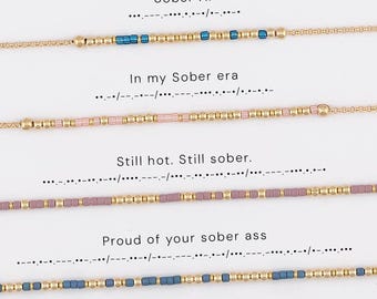 May include: Four gold-toned chain bracelets featuring Morse code patterns. The first has blue beads, the second light pink, the third mauve, and the fourth slate blue. Each bracelet is displayed below its corresponding text: Sober AF, In my Sober era, Still hot. Still sober., and Proud of your sober ass.