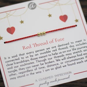 Könnte beinhalten: Ein rotes Armband mit drei goldenen Perlen, präsentiert auf einer weißen Karte mit dem Text "Red Thread of Fate". Die Karte zeigt rote Herzen und goldene Sterne, mit einer Botschaft über Verbindungen und Seelenverwandte.