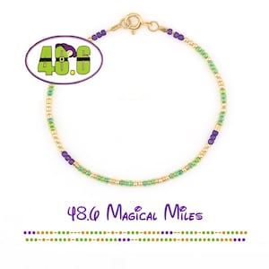 Puede incluir: Una delicada pulsera de cuentas doradas con detalles verdes y morados. La pulsera tiene un cierre y está adornada con pequeñas cuentas de colores. La imagen incluye el texto "48.6 Magical Miles" en una fuente caprichosa.