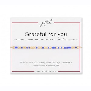 May include: A delicate bracelet featuring alternating small gold-tone beads and vibrant blue cylindrical glass beads, displayed on a white card with the text "gifted.", "Grateful for you", Morse code, and "14k Gold Fill or .925 Sterling Silver".