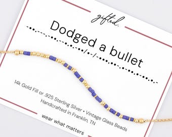 May include: A gold chain bracelet featuring alternating purple and gold beads arranged in a Morse code pattern. The bracelet is displayed on a white card with the text "gifted. Dodged a bullet" and "14k Gold Fill or .925 Sterling Silver • Vintage Glass Beads Handcrafted in Franklin, TN wear what matters".