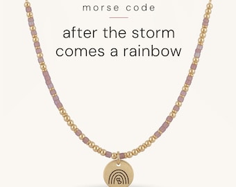 May include: A gold chain necklace featuring alternating metallic gold and matte mauve beads, with a central circular gold pendant engraved with a rainbow and two small hearts. The text "morse code after the storm comes a rainbow" is visible.