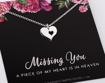 May include: A silver heart pendant necklace featuring a small heart-shaped cutout, displayed on a black card with pink floral accents. The card features the text "Missing You" and "A PIECE OF MY HEART IS IN HEAVEN" along with a website address.