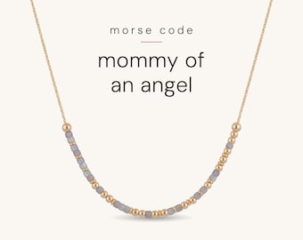 May include: A gold chain necklace featuring a Morse code pattern made of small metallic gold beads and matte grey beads. The text "morse code mommy of an angel" is printed above the necklace in a simple, dark grey sans-serif font.