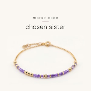 May include: A gold chain bracelet featuring a Morse code pattern made of small purple cylindrical beads and round gold beads. The text "morse code" and "chosen sister" is printed above the bracelet. It includes a gold clasp and a small circular tag.