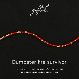 May include: Red beaded necklace with gold accents. The text "gifted" is written in cursive at the top. The phrase "Dumpster fire survivor" is written in a bold, sans-serif font below the necklace.
