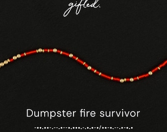 May include: A beaded necklace featuring alternating red cylindrical beads and small gold spherical beads. The text "gifted." appears at the top, while the text "Dumpster fire survivor" and Morse code are printed at the bottom.