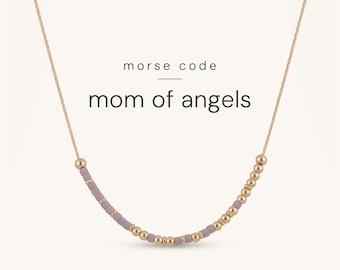 May include: A delicate gold chain necklace featuring a Morse code pattern made of small, round gold-tone beads and cylindrical mauve-coloured beads. The text "morse code" and "mom of angels" is printed above the necklace in black font.