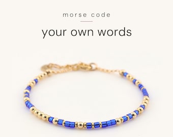 May include: A delicate bracelet featuring alternating metallic gold-tone beads and vibrant blue cylindrical beads arranged in a Morse code pattern. The text "morse code" and "your own words" is printed above the jewellery piece.