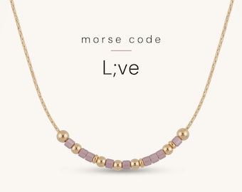 May include: A gold-toned chain necklace featuring a central segment of alternating small round gold beads and cylindrical mauve beads arranged in a Morse code pattern. The text "morse code" and "L;ve" is printed above the necklace.