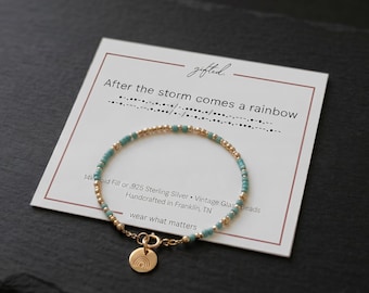 May include: A delicate bracelet featuring alternating turquoise and gold-toned beads on a display card with the text After the storm comes a rainbow and Morse code. A small circular charm with a rainbow engraving hangs from the gold-toned clasp.