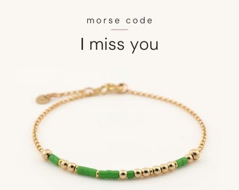 May include: A gold-toned chain bracelet featuring a Morse code pattern made of small, cylindrical green beads and round gold-toned beads. The text "morse code" and "I miss you" is printed above the bracelet on a plain white background.