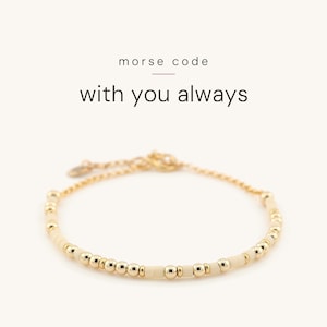 May include: A gold-toned chain bracelet featuring alternating small metallic gold beads and matte cream-colored beads arranged in a pattern. The text above the bracelet reads morse code with you always in black lettering.
