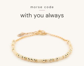 May include: A gold-toned chain bracelet featuring alternating small metallic gold beads and matte cream-coloured beads arranged in a pattern. The text above the bracelet reads morse code with you always in black lettering.