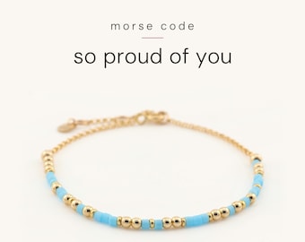 May include: A gold-toned chain bracelet featuring a pattern of small, round gold beads and light blue cylindrical beads arranged in Morse code. The text above the bracelet reads morse code so proud of you.