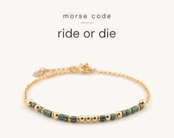 May include: A gold chain bracelet featuring a Morse code pattern made of small, cylindrical dark green beads and round gold beads. The text "morse code" and "ride or die" is printed above the bracelet in a simple black font.