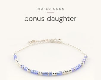 May include: A silver chain bracelet featuring a pattern of small blue and silver beads arranged in Morse code. The text above the bracelet reads morse code bonus daughter. The bracelet has a delicate silver chain with a clasp closure.