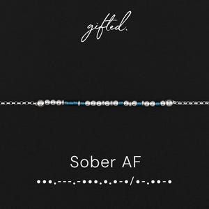 May include: A silver-toned beaded bracelet with a dark blue center section. The bracelet features the text "Sober AF" in white, along with a Morse code pattern. The word "gifted" is written in cursive at the top.