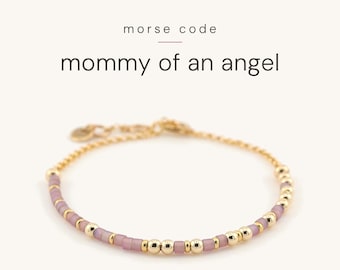 May include: A gold-toned chain bracelet featuring a pattern of small, matte lavender beads and polished gold-coloured beads arranged in Morse code. The text "morse code" and "mommy of an angel" is printed in black above the bracelet.